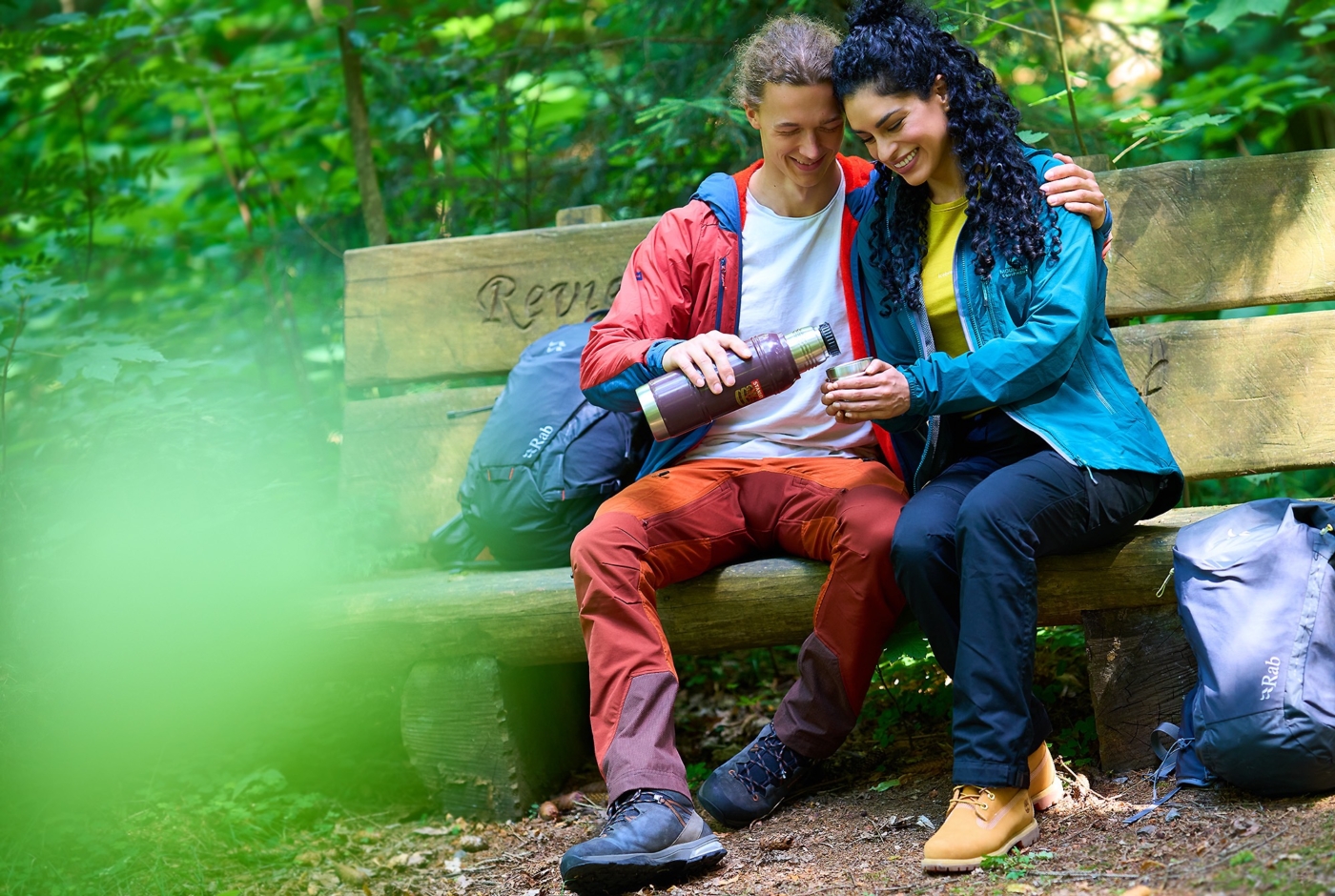 Glückliches Paar im Wanderoutfit sitzend auf einer Bank im Wald bei einer Rast. Mann schenkt frischen Tee aus