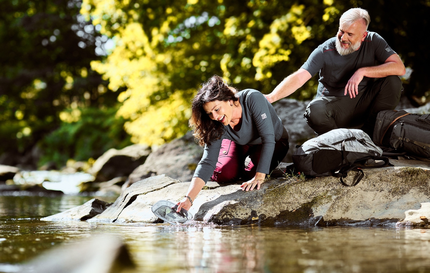Glückliches Paar im Wanderoutfit bei einer Rast ihrer Wanderung am Fluss Wasser schöpfend