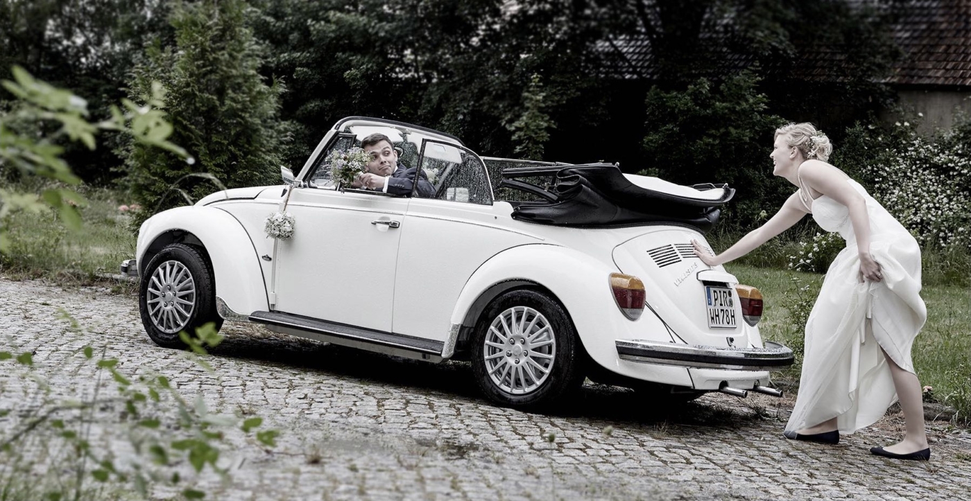 Die Braut in weiss schiebt das Auto des Hochzeitspaares an während der Bräutigam mit Brautstrauss im auto sitzt