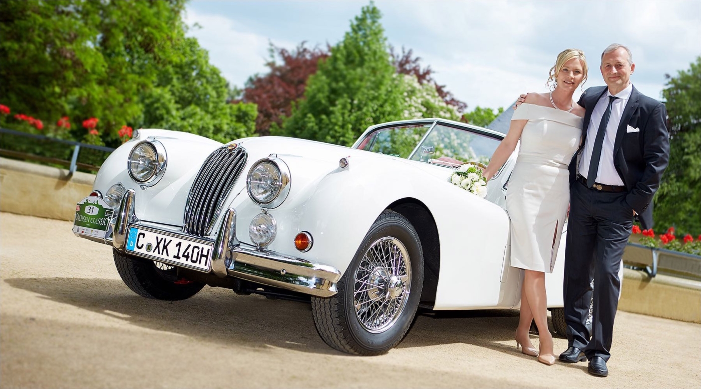 Brautpaar steht locker umarmt neben ihrem weissen Oldtimer