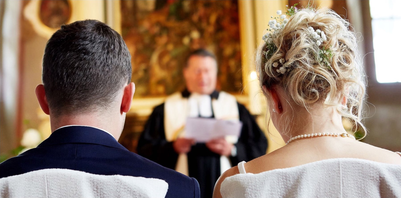 Rückenportrait eines Brautpaars. Im Hintergrund der Pfarrer unscharf während der Trauzeremonie.