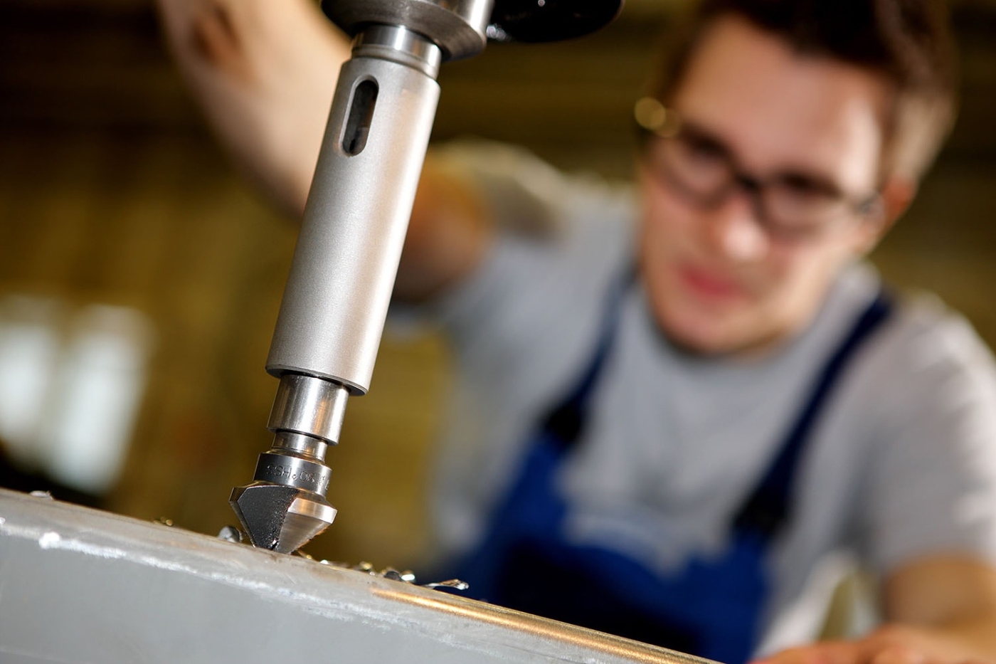 Azubi mit Schutzbrille unscharf im Hintergrund beim Bohren eines Werkstückes