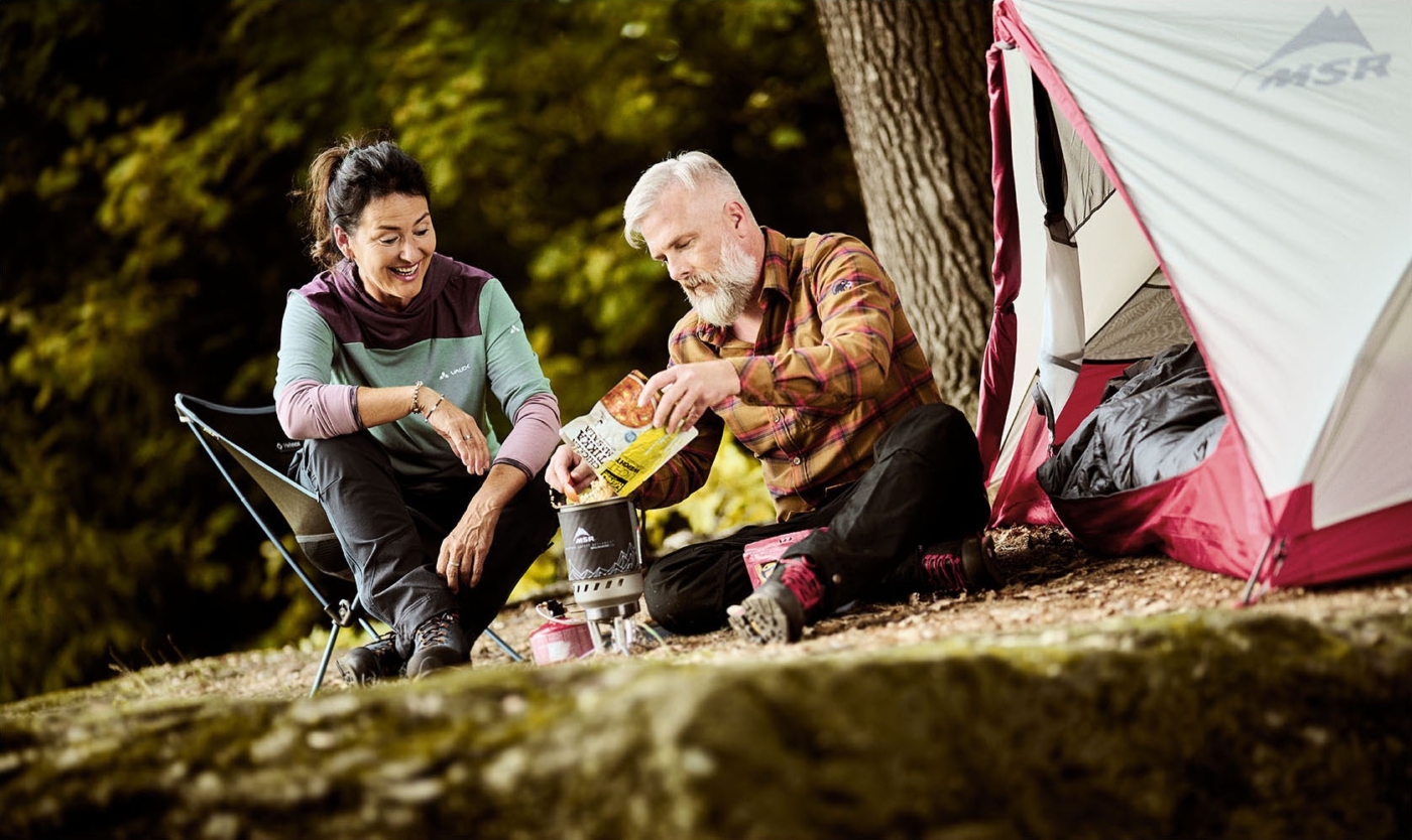 Mann und Frau vor Zelt sitzend. Mann bereitet eine Suppe auf einem Campingkocher zu