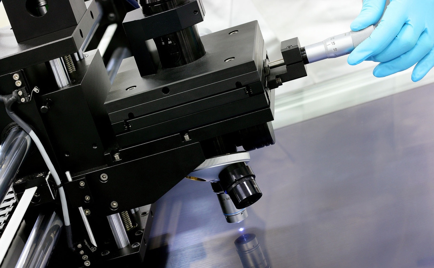 Mikroskopische Vermessung einer Metalloberfläche. Hand mit blauem Handschuh bedient eine Messschraube