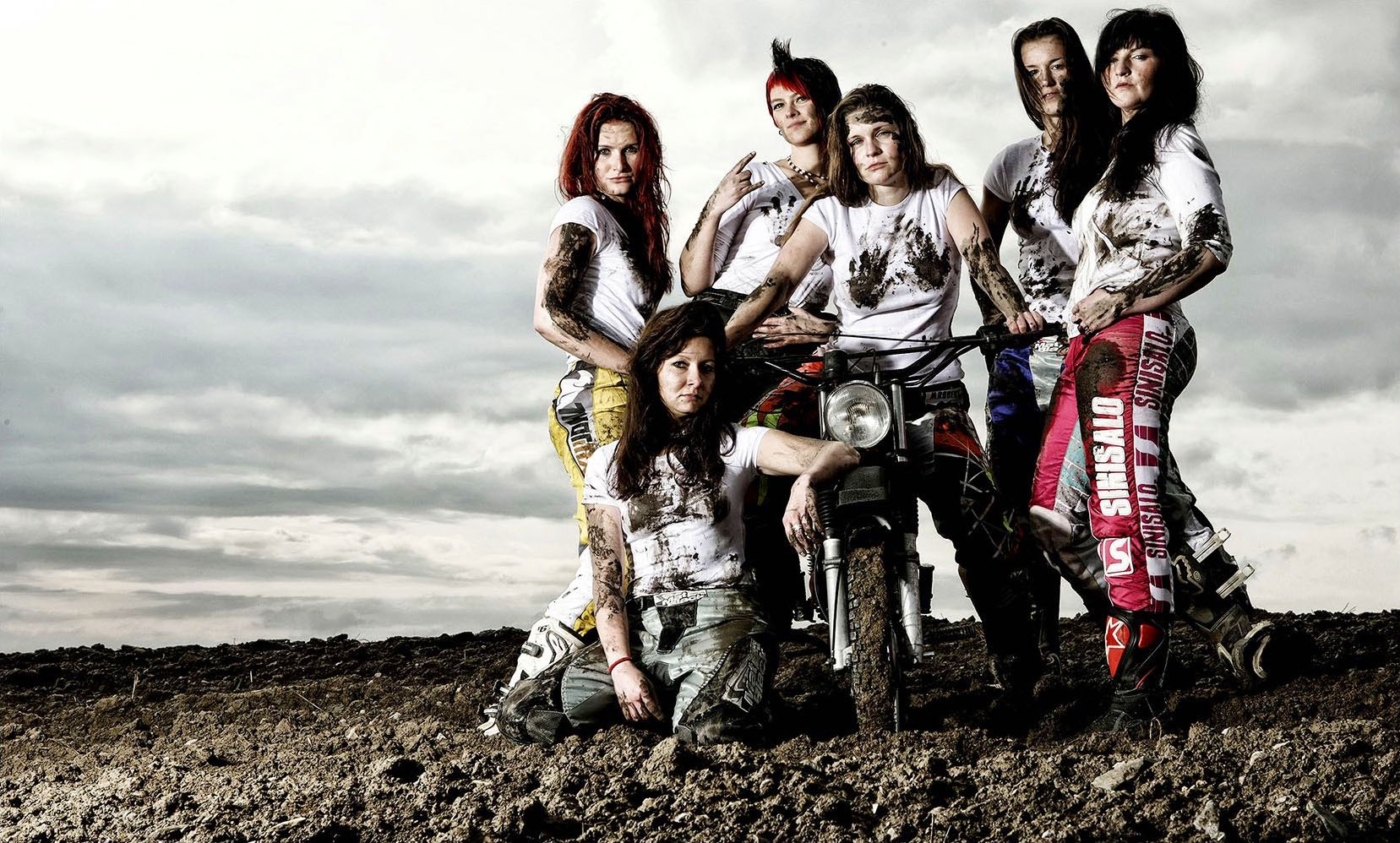 Mopedgang. 6 Frauen in dreckiger Motorradbekleidung an einem Motorrad gelehnt auf einem braunen Stoppelfeld