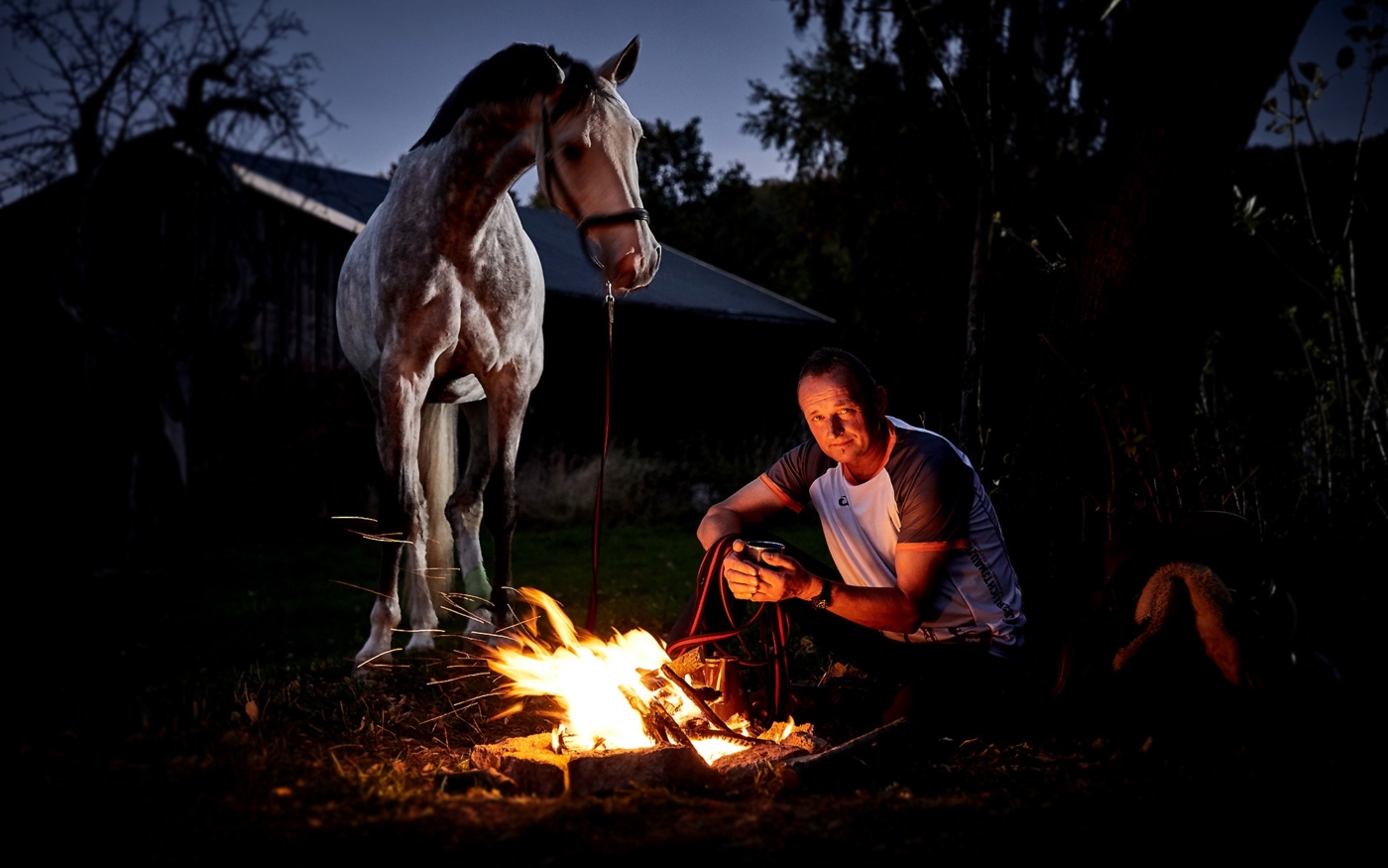 Pferd mit Besitzer der am Lagerfeuer sitzt und Kaffee trinkt in der blauen Stunde